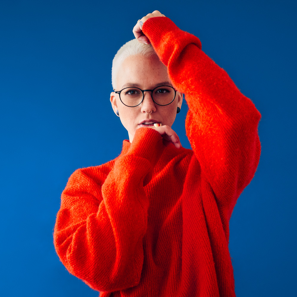 Stefanie Heinzmann Portrait mit orangen Pullover vor blauem Background.